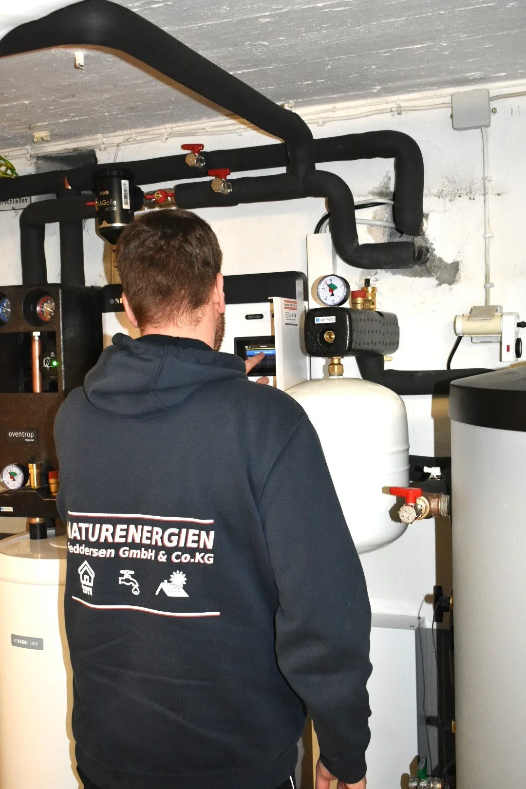 Monteur im dunklen Hoodie mit Aufdruck Naturenergien Feddersen GmbH & Co.KG in einem Heizungsraum