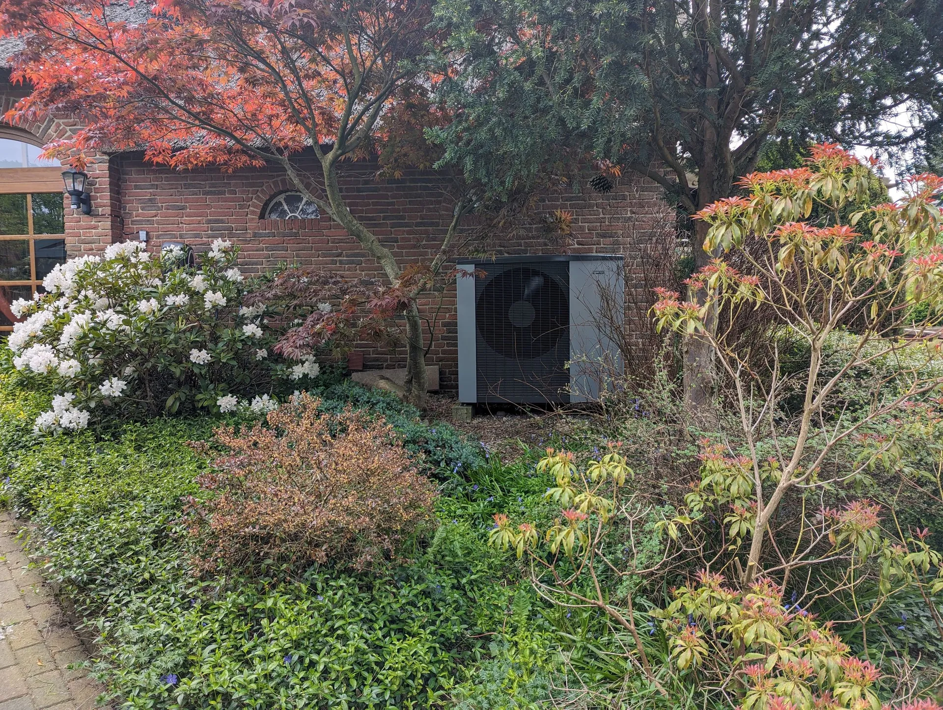 Wärmepumpe im Vorgarten vor einer Klinkerwand, umgeben von Rhododendren und Ahorn