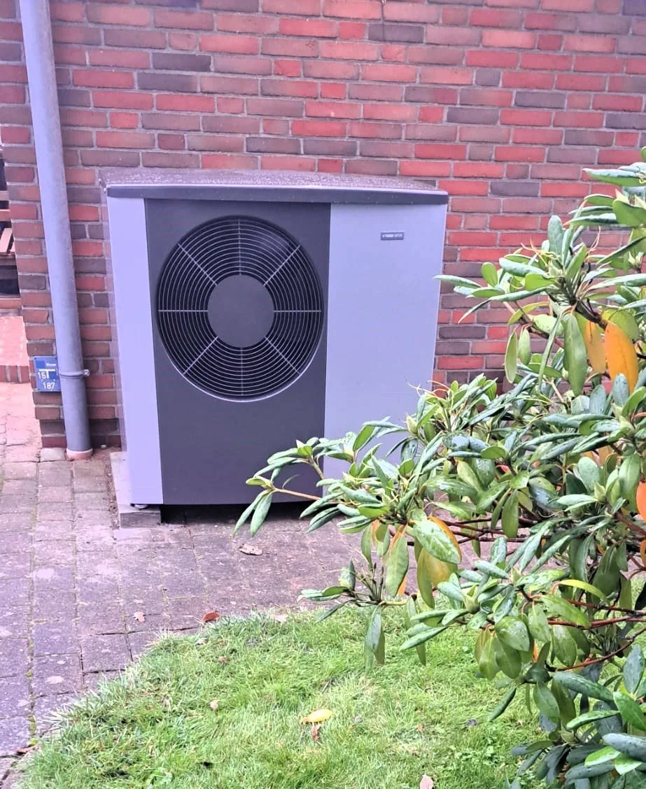 Wärmepumpe an einem roten Klinkerhaus neben einem Strauch