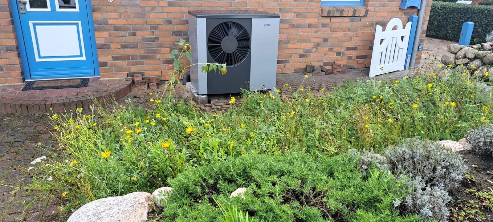 Wärmepumpe vor einem Klinkerhaus mit blauer Tür und Blumenbeet